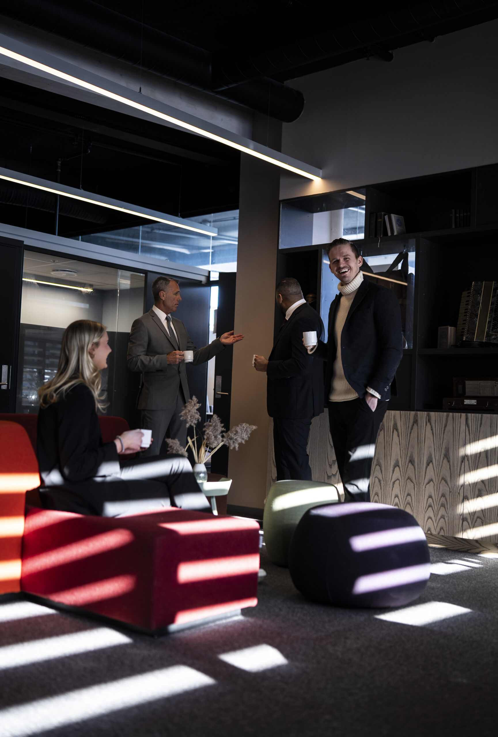 Flere kollegaer i dress står og sitter i et moderne loungeområde med fargerike møbler og kaffekopper i hånden.